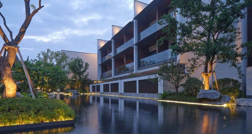A modern building with three stories, featuring balconies and large glass doors, is surrounded by greenery and illuminated at dusk. A calm, reflective water feature runs alongside the structure, bordered by trees, shrubs, and accent lighting, creating a serene and elegant atmosphere.